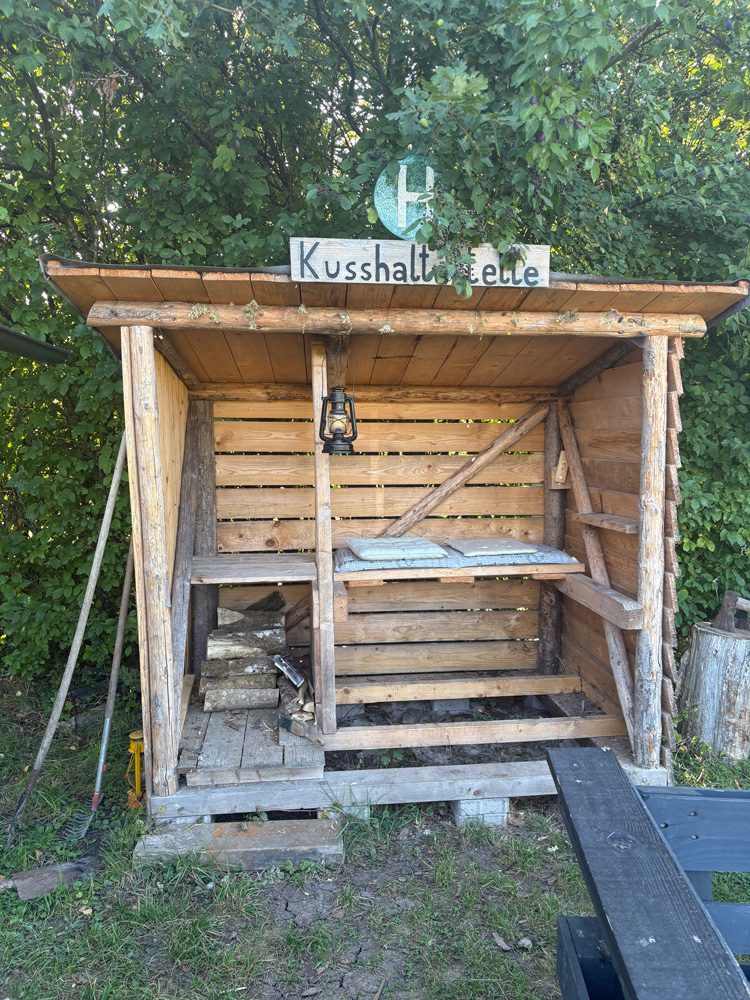 Holzunterstand mit Schild „Kusshaltestelle“ im Grünen.