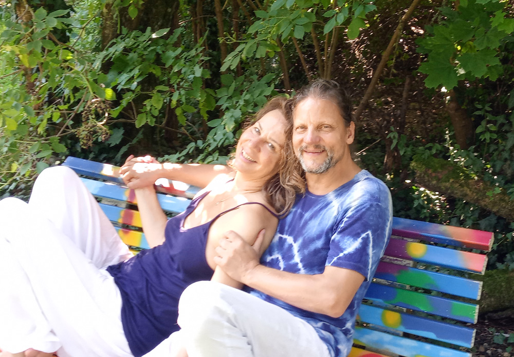 Lächelnde Martina und Christian sitzen nah beieinander auf bunter Gartenbank.