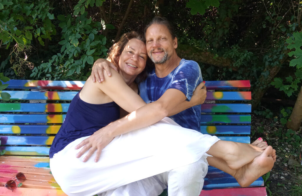 Zufriedene Martina und Christian umarmen sich auf bunter Bank im Grünen, symbolisch für Verbindung und Nähe.